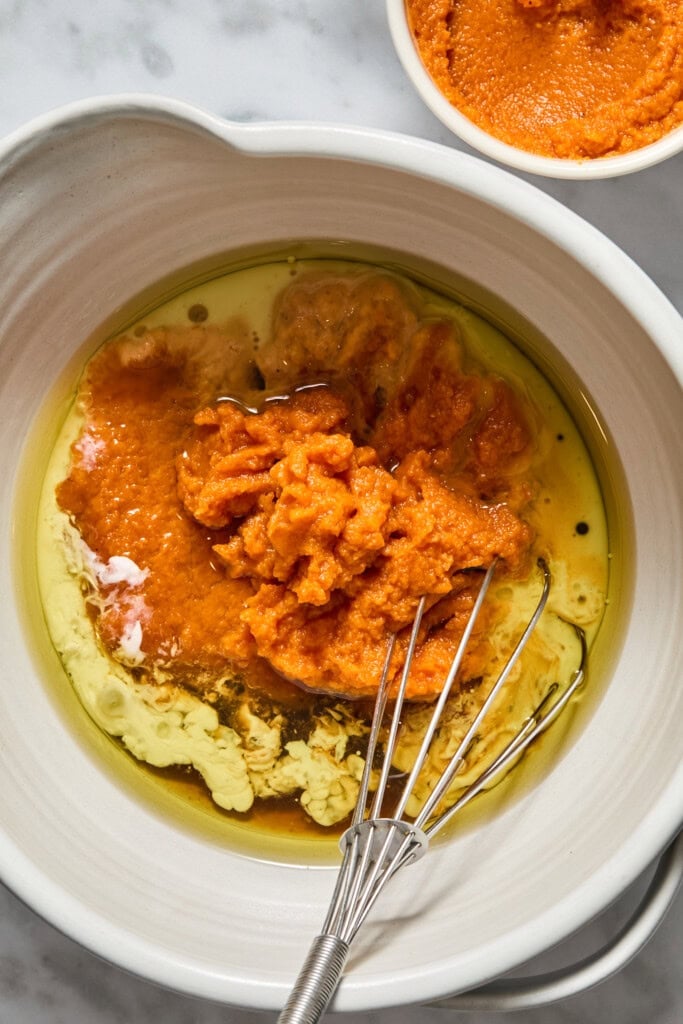 A mixing bowl with pumpkin puree, oil, and liquid ingredients for Vegan Pumpkin Bread sits next to a whisk and a small bowl of pumpkin puree on a marble surface.