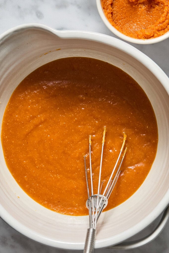 A white mixing bowl filled with orange pumpkin puree mixture and a metal whisk, perfect for making Vegan Pumpkin Bread. A small bowl of more pumpkin puree sits nearby on a marble countertop.