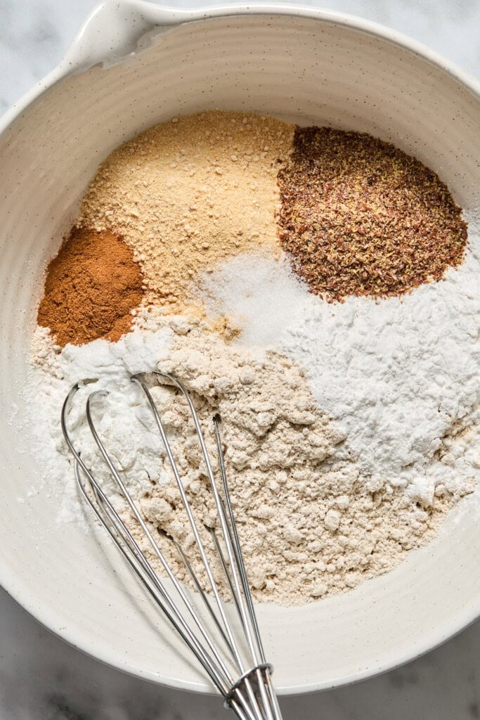 A mixing bowl with various dry baking ingredients, including flour, flaxseed, cinnamon, powder, and sugar—perfectly prepped for making delicious Vegan Pumpkin Bread—with a metal whisk resting inside.
