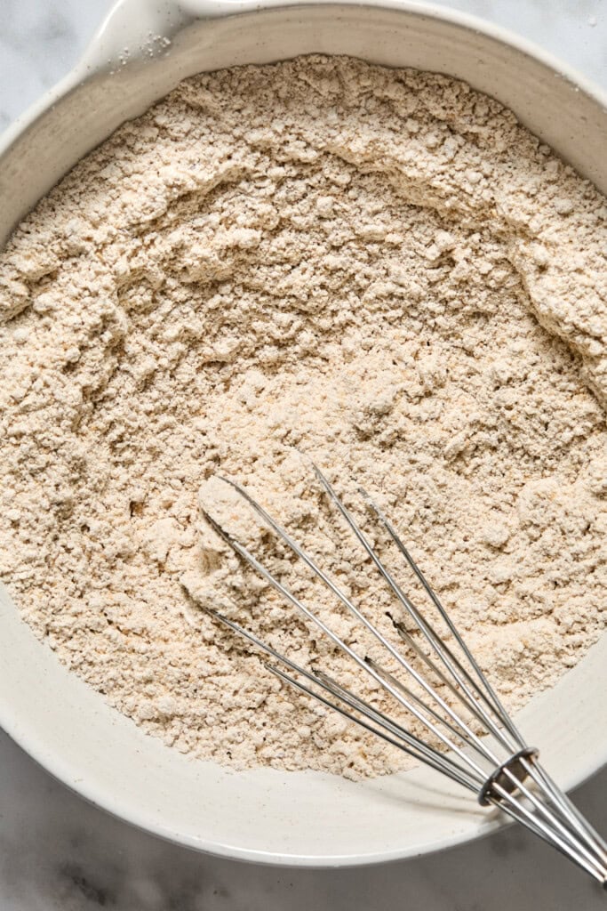 A metal whisk rests in a bowl filled with a light beige, crumbly dry mixture—likely the flour and spices for Vegan Pumpkin Bread—on a marble surface.