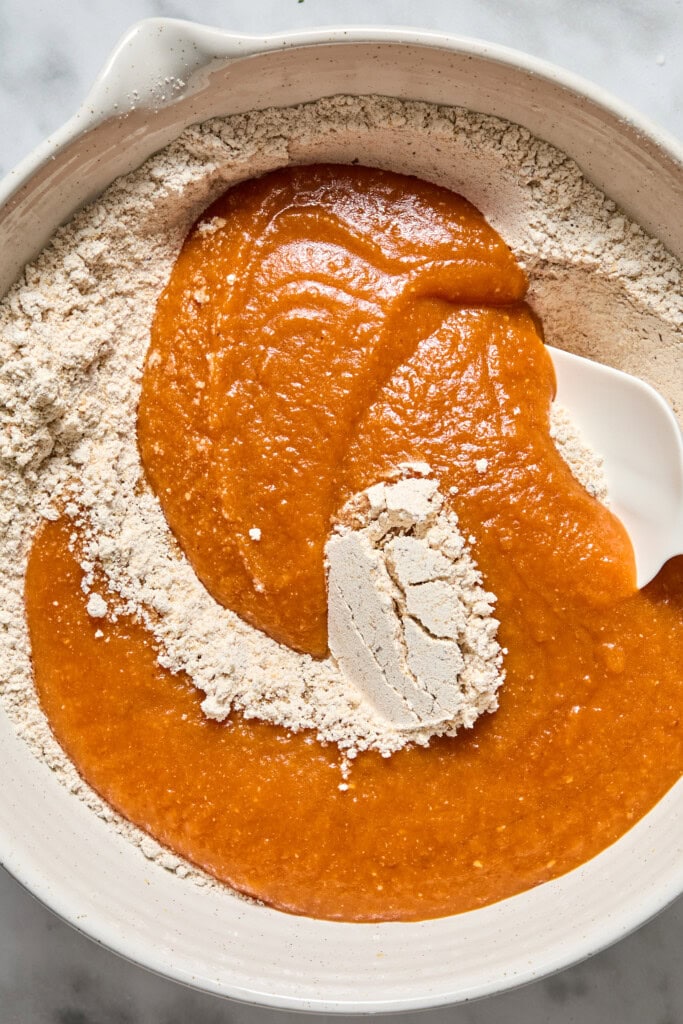 A mixing bowl with dry flour mixture and orange pumpkin puree being combined with a white spatula, ready for baking delicious Vegan Pumpkin Bread.