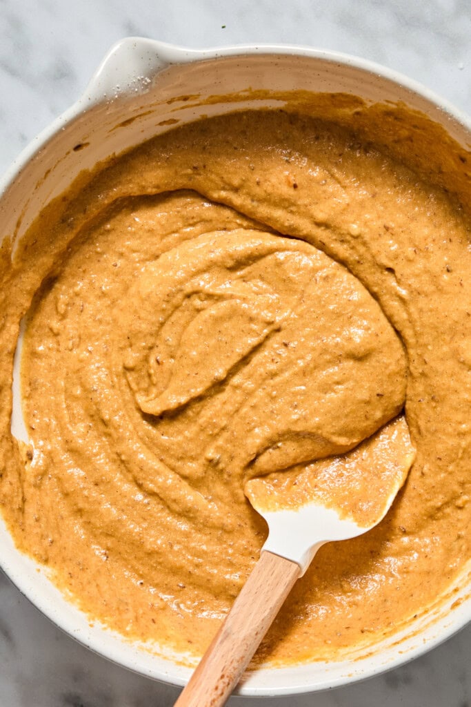 A white mixing bowl filled with thick, orange-brown Vegan Pumpkin Bread batter and a white spatula with a wooden handle resting inside.