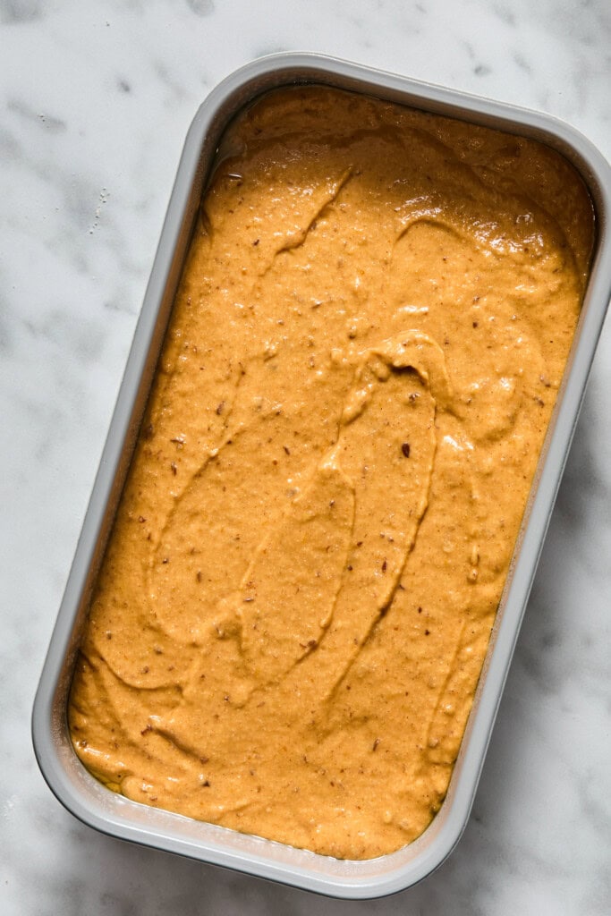 A rectangular dish filled with a smooth, orange-brown Vegan Pumpkin Bread batter sits on a white marble surface, ready to be baked or frozen.