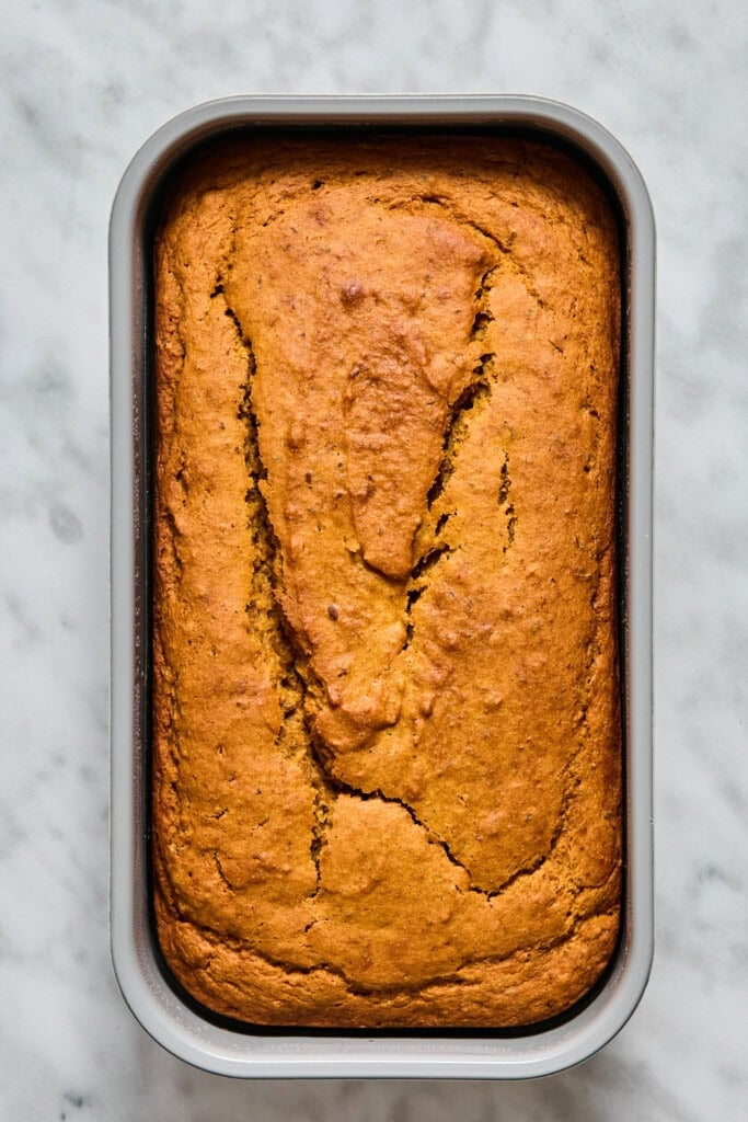 A baked loaf of Vegan Pumpkin Bread with a cracked top sits in a rectangular metal pan on a marble surface.