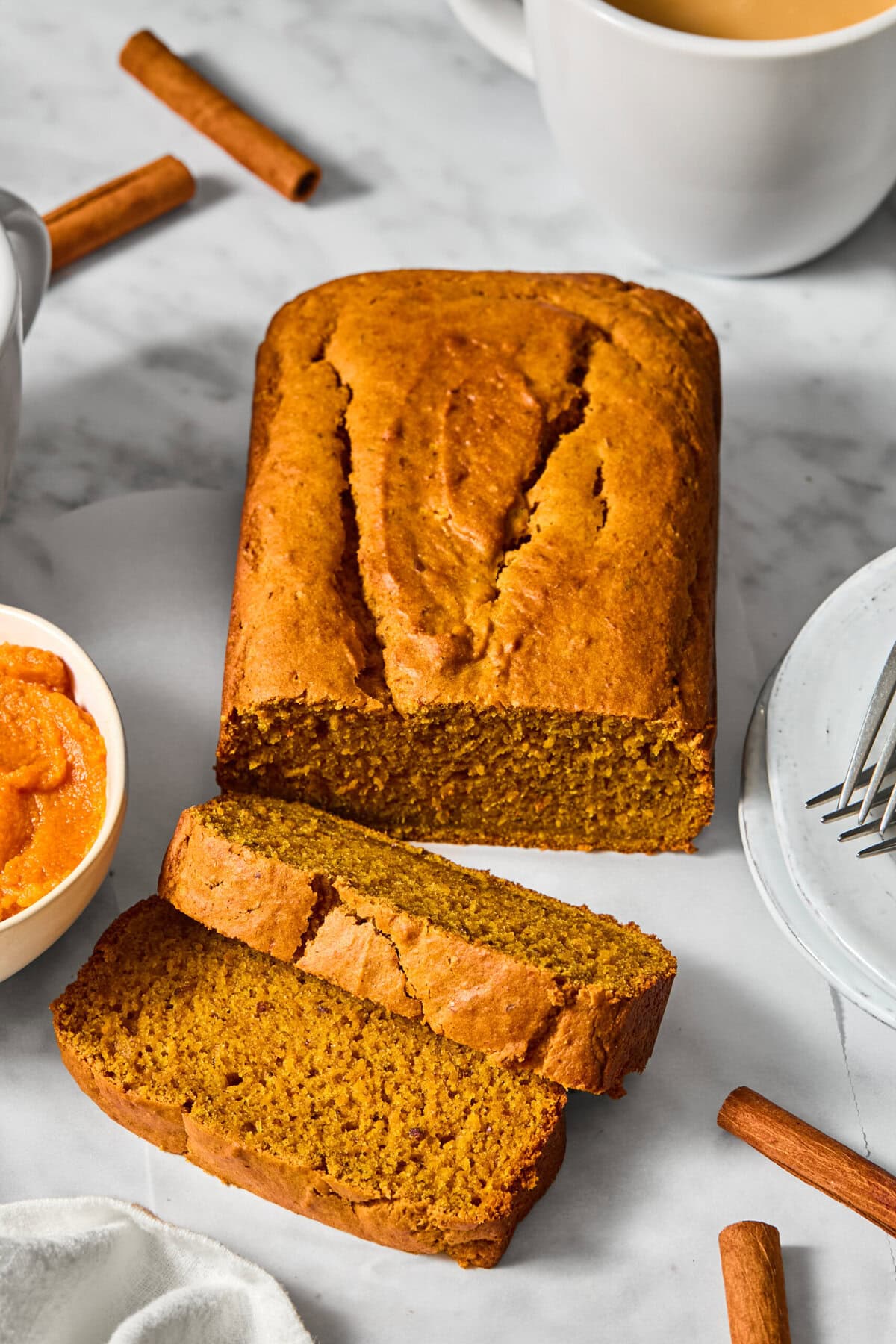 A loaf of Vegan Pumpkin Bread with two slices cut, surrounded by cinnamon sticks, a bowl of pumpkin puree, a cup of coffee, and plates with forks on a marble surface.