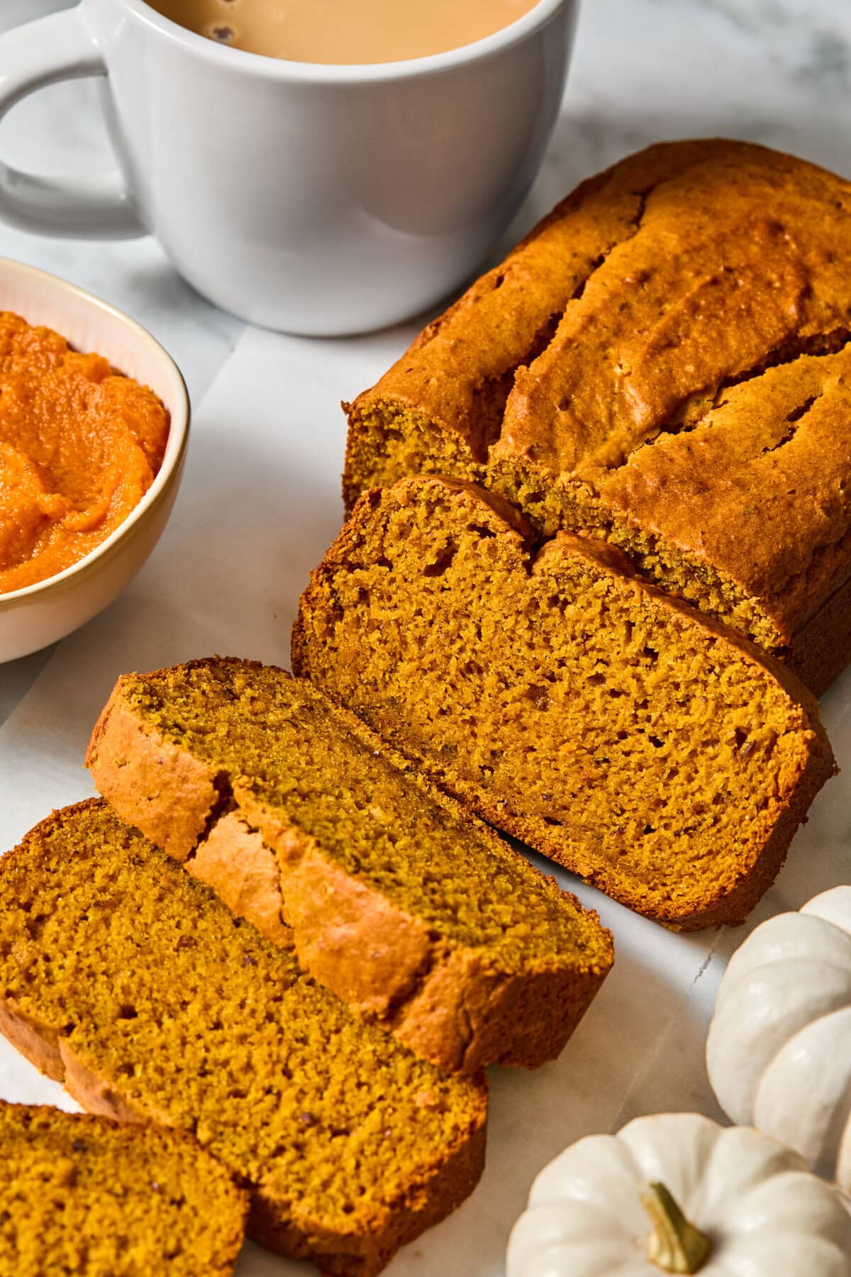 Vegan Pumpkin Bread is sliced on parchment paper next to a white mug of coffee, a bowl of pumpkin puree, and small white pumpkins on a white surface.