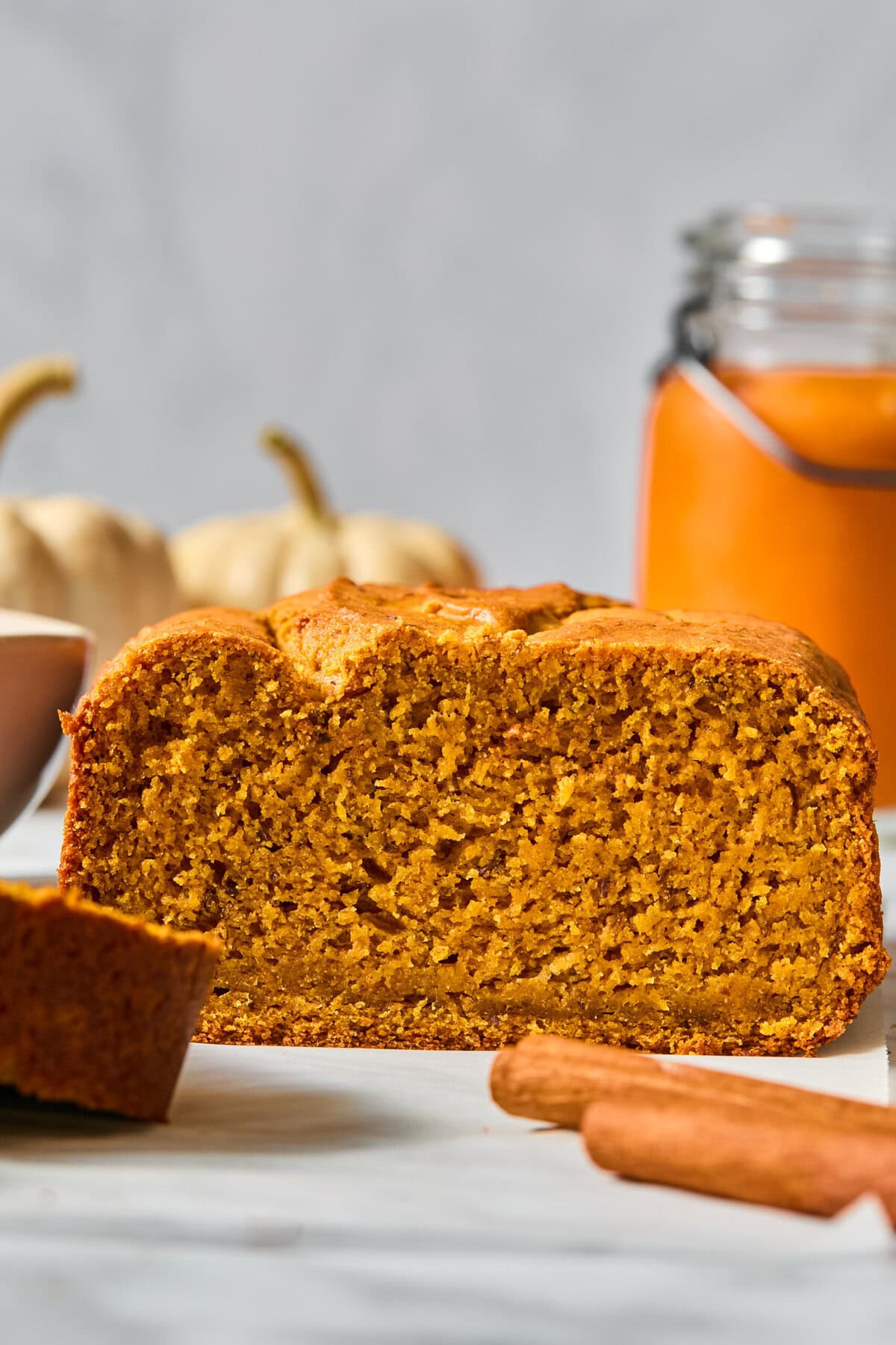 A sliced loaf of Vegan Pumpkin Bread sits on a white surface, with cinnamon sticks and a jar of orange spread in the background. Two small pumpkins are also visible, giving an autumnal feel.