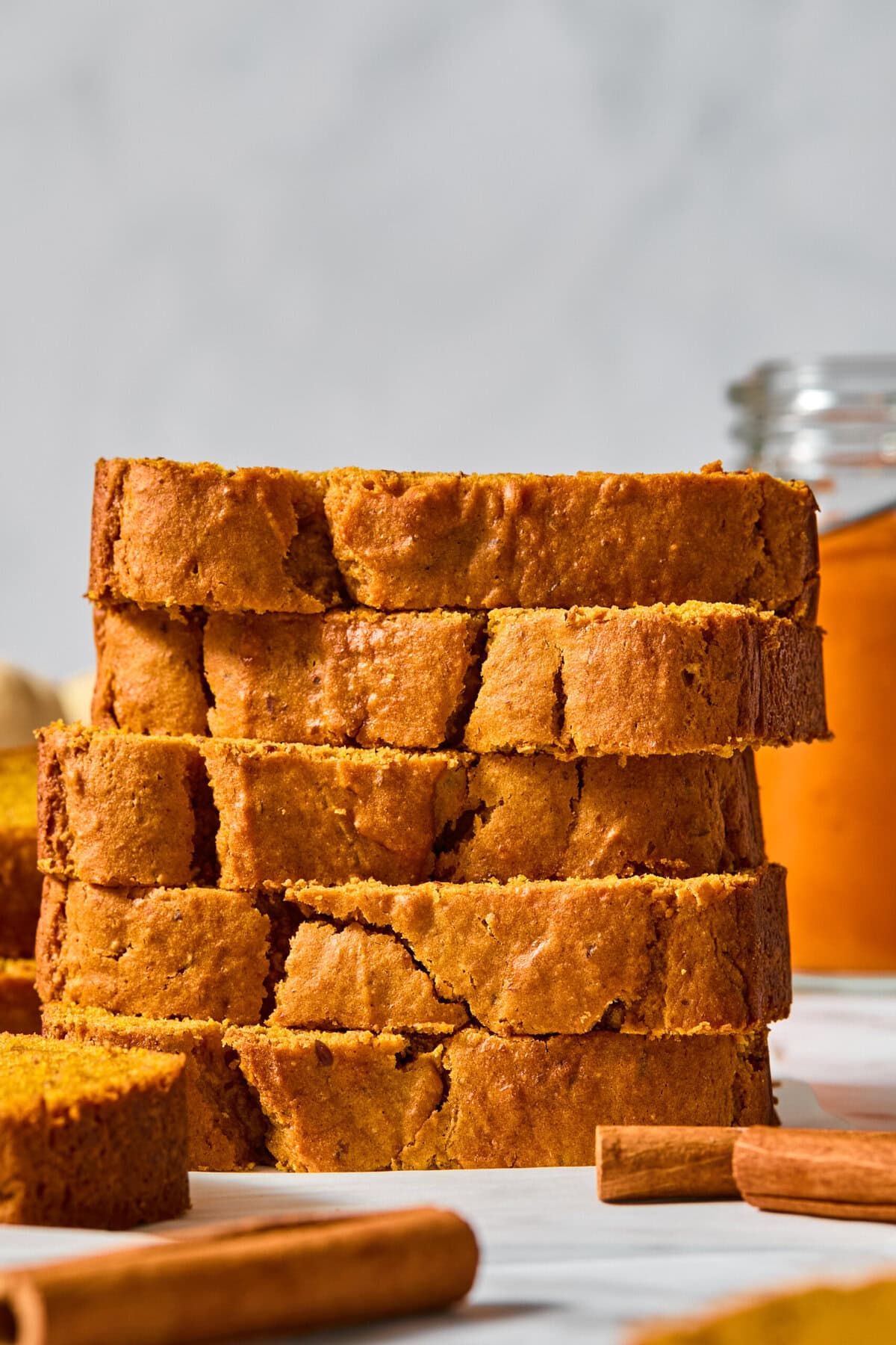 A stack of sliced vegan pumpkin bread sits on a white surface with cinnamon sticks nearby and a jar of orange spread in the background.