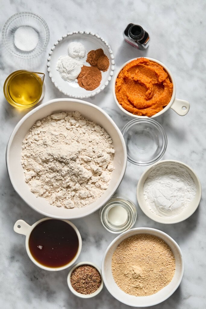 Bowls of flour, sugar, pumpkin puree, oil, spices, vanilla, and various baking ingredients are arranged on a marble countertop, prepared for baking a delicious Vegan Pumpkin Bread.