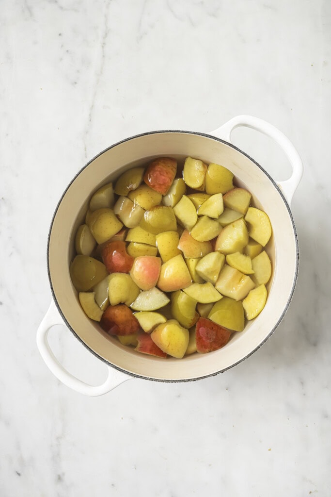A white pot filled with chopped apples, some red and some yellow, sits on a marble countertop. The apples are partially submerged in liquid, ready to be transformed into delicious homemade Instant Pot applesauce.