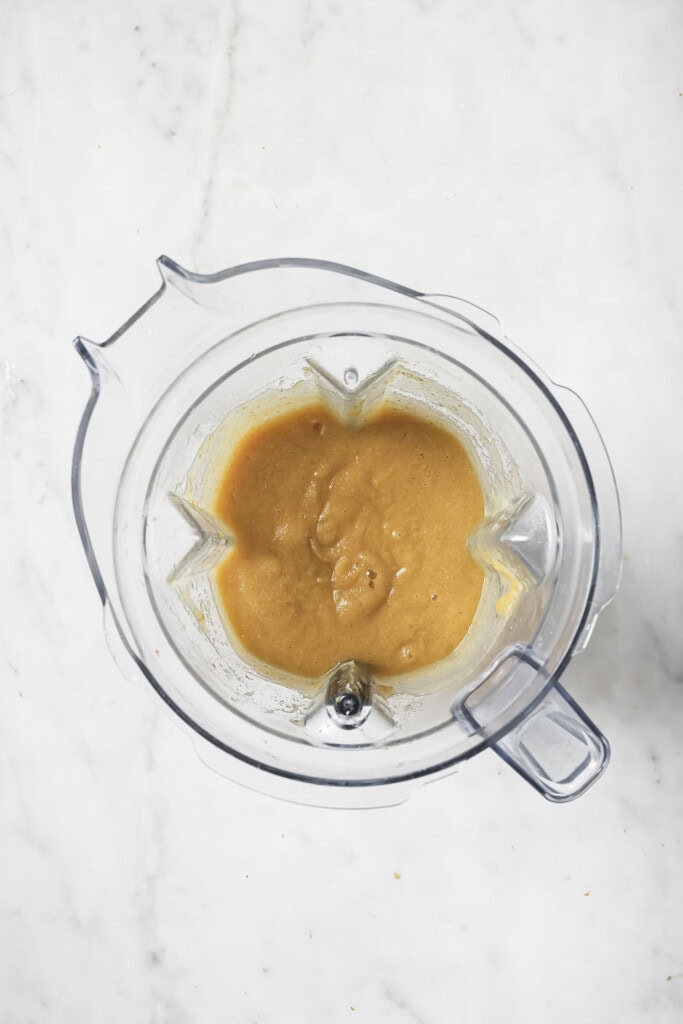 Top view of a blender with creamy, light brown Homemade Instant Pot Applesauce inside, placed on a white marble surface.