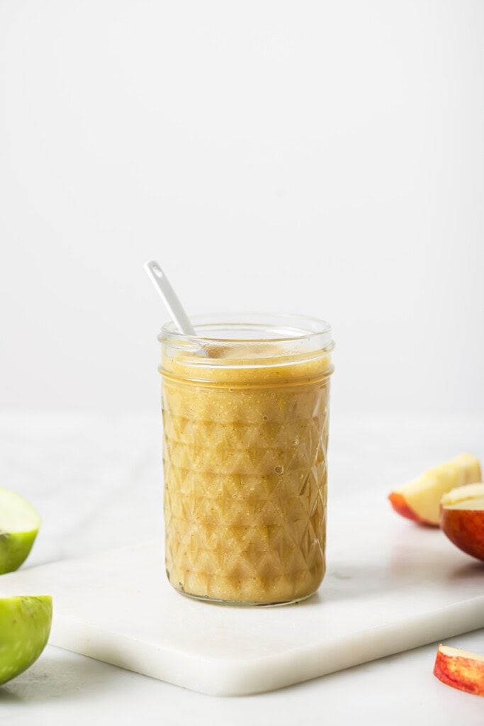 A glass jar filled with homemade applesauce and a spoon inside, placed on a white surface with sliced green and red apples nearby.