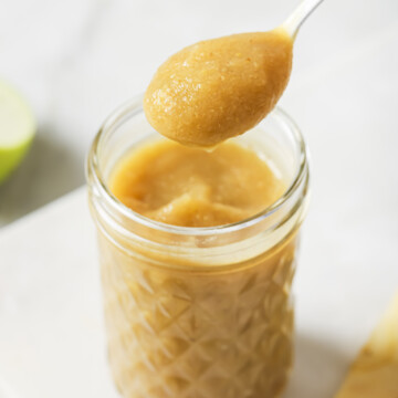 A spoonful of homemade applesauce is held above a glass jar filled with applesauce, with apple slices blurred in the background.
