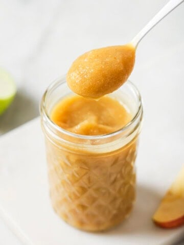 A spoonful of homemade applesauce is held above a glass jar filled with applesauce, with apple slices blurred in the background.
