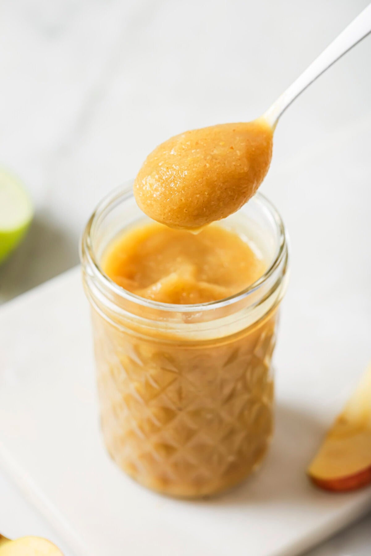 A spoonful of homemade applesauce is held above a glass jar filled with applesauce, with apple slices blurred in the background.