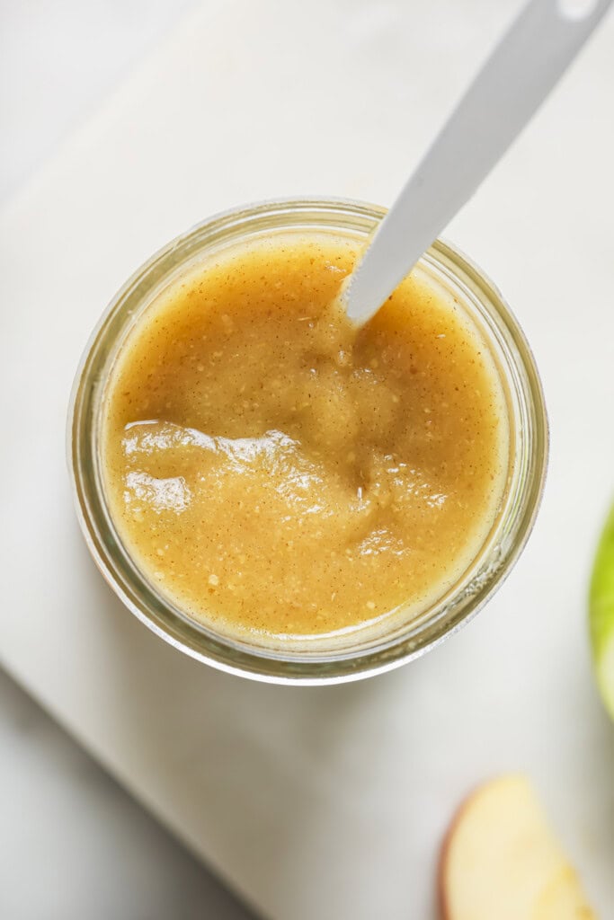 A glass jar filled with smooth, light brown homemade applesauce and a white spoon inside. Part of a sliced apple is visible in the corner on a white surface.