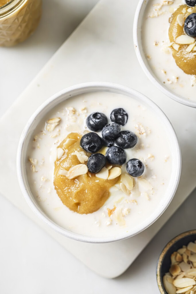 A bowl of oatmeal topped with blueberries, almond slices, and a swirl of homemade applesauce, sitting on a white surface.