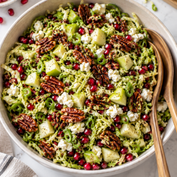 A bowl of shredded brussels sprouts salad with apple chunks, pecans, pomegranate seeds, green onions, and crumbled cheese, with serving utensils on the side.