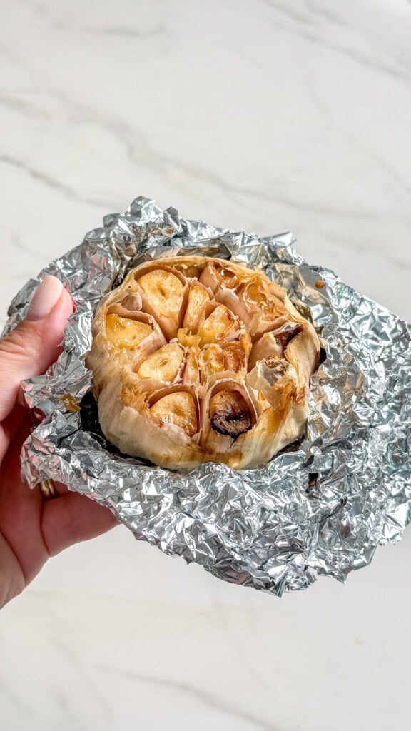 A hand holding a head of roasted garlic wrapped in aluminum foil, a key ingredient for vegan mashed cauliflower.