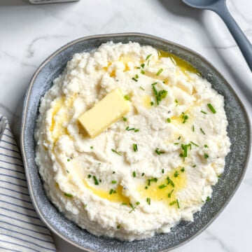 A bowl of mashed cauliflower topped with a pat of butter, melted butter, and chopped chives, with a salt shaker and spoon nearby; an easy mashed cauliflower recipe.