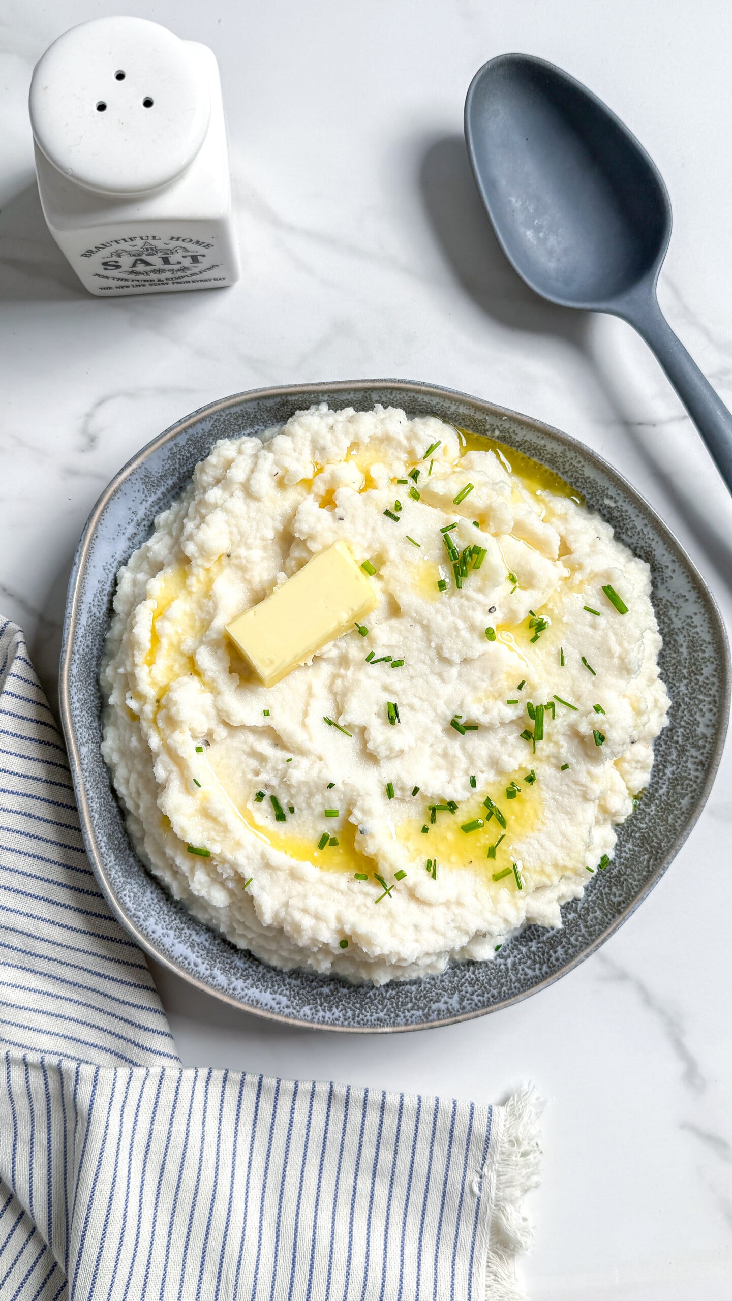 A bowl of mashed cauliflower topped with a pat of butter, melted butter, and chopped chives, with a salt shaker and spoon nearby; an easy mashed cauliflower recipe.