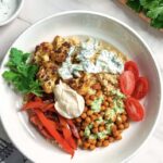 A bowl filled with quinoa, roasted cauliflower, chickpeas, red bell peppers, sliced cherry tomatoes, fresh parsley, and dollops of green and white sauces—these Vegan Shawarma Bowls are served on a white countertop with herbs and sauce in the background.