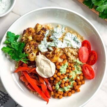 A bowl filled with quinoa, roasted cauliflower, chickpeas, red bell peppers, sliced cherry tomatoes, fresh parsley, and dollops of green and white sauces—these Vegan Shawarma Bowls are served on a white countertop with herbs and sauce in the background.