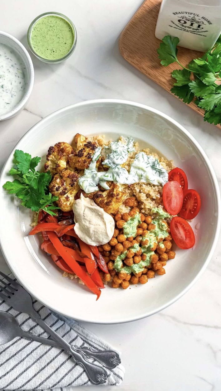 A bowl filled with quinoa, roasted cauliflower, chickpeas, red bell peppers, sliced cherry tomatoes, fresh parsley, and dollops of green and white sauces—these Vegan Shawarma Bowls are served on a white countertop with herbs and sauce in the background.