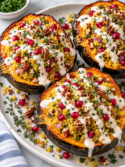 Three roasted acorn squash halves filled with couscous, red onion, pecans, pomegranate seeds, fresh herbs, and drizzled with a creamy white sauce are served on a white platter. This visually stunning stuffed acorn squash makes for a delicious centerpiece. A small bowl of herbs is in the background.