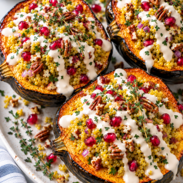 Three roasted acorn squash halves filled with couscous, red onion, pecans, pomegranate seeds, fresh herbs, and drizzled with a creamy white sauce are served on a white platter. This visually stunning stuffed acorn squash makes for a delicious centerpiece. A small bowl of herbs is in the background.