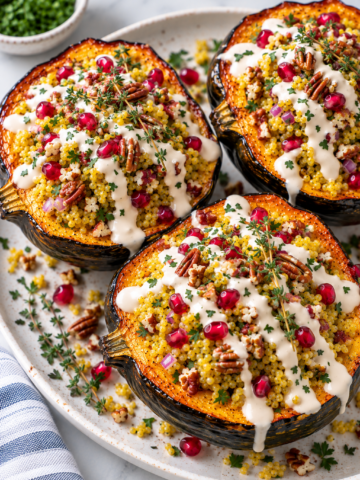 Three roasted acorn squash halves filled with couscous, red onion, pecans, pomegranate seeds, fresh herbs, and drizzled with a creamy white sauce are served on a white platter. This visually stunning stuffed acorn squash makes for a delicious centerpiece. A small bowl of herbs is in the background.