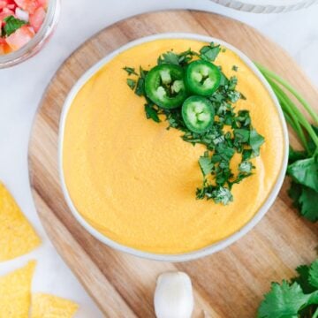 vegan nacho cheese topped with sliced jalapeno and cilantro in a white bowl on a brown cutting board with nachos, cilantro and salsa on the side