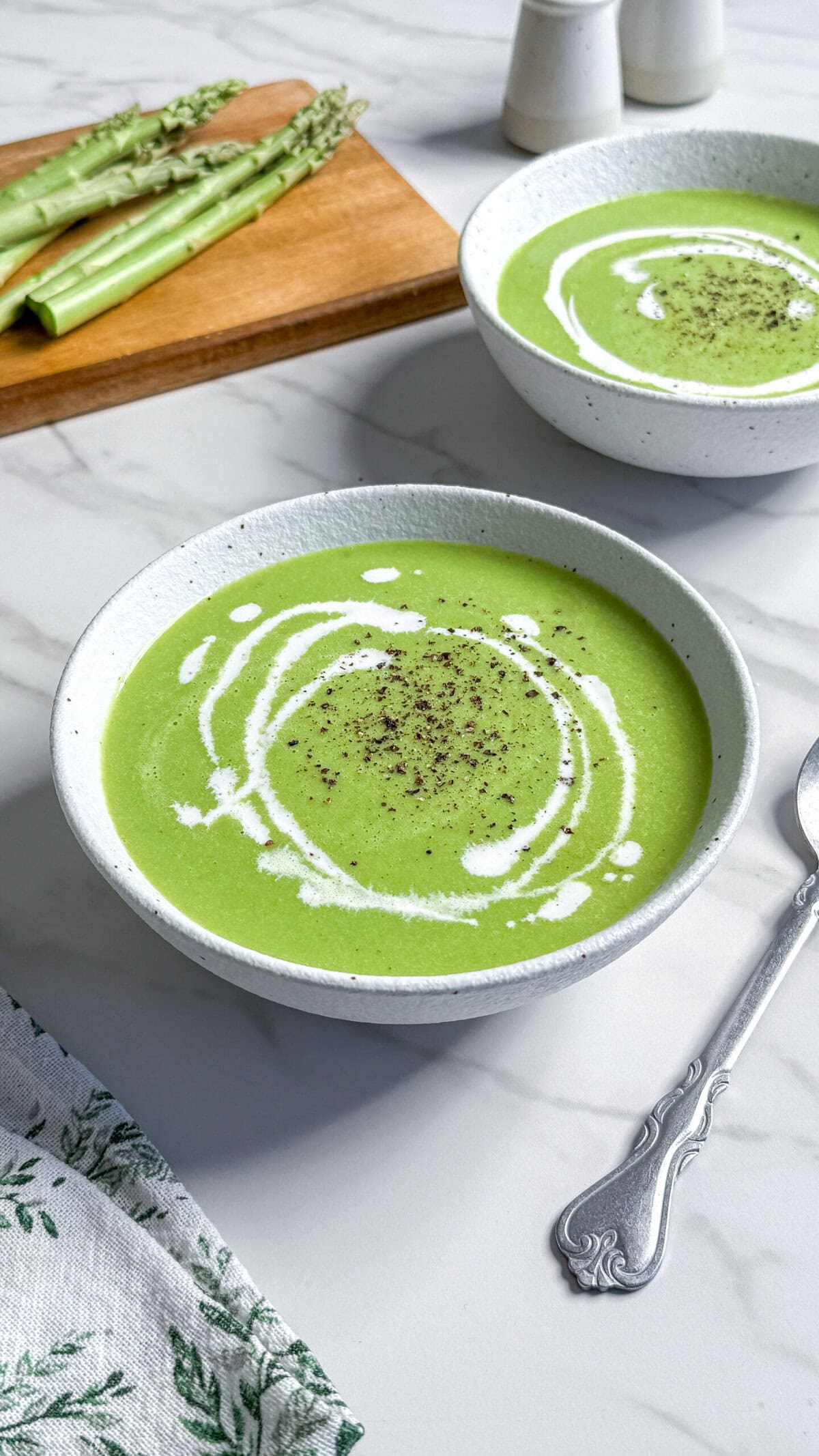 Two bowls of vegan cream of asparagus soup, garnished with a swirl of cream and cracked black pepper, sit on a marble countertop. A bunch of fresh asparagus spears rests on a wooden board in the background.