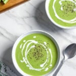 Two bowls of vegan cream of asparagus soup topped with a swirl of cream and black pepper sit on a white marble surface, next to a spoon, a patterned napkin, and fresh asparagus on a wooden board.