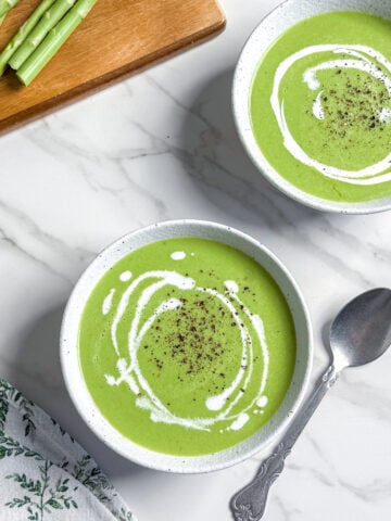 Two bowls of vegan cream of asparagus soup topped with a swirl of cream and black pepper sit on a white marble surface, next to a spoon, a patterned napkin, and fresh asparagus on a wooden board.