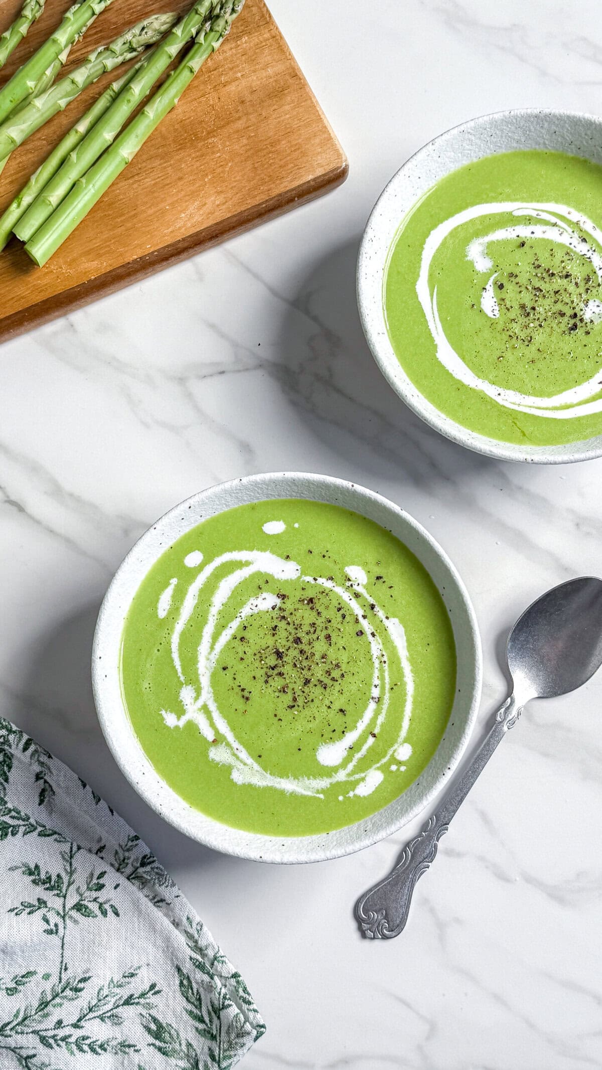 Two bowls of vegan cream of asparagus soup topped with a swirl of cream and black pepper sit on a white marble surface, next to a spoon, a patterned napkin, and fresh asparagus on a wooden board.