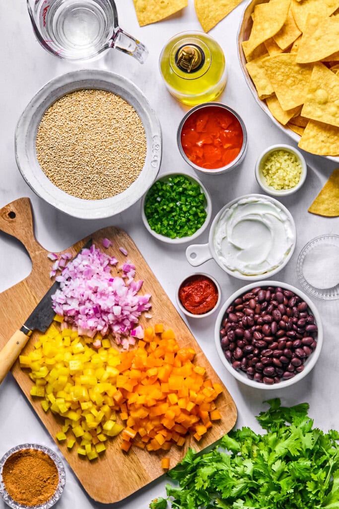 Chopped yellow and orange bell peppers, red onion, black beans, jalapeños, diced tomatoes, garlic, sour cream, quinoa, olive oil, spices, cilantro, and tortilla chips arranged on a white surface—perfect for assembling a flavorful Vegan Mexican Casserole.