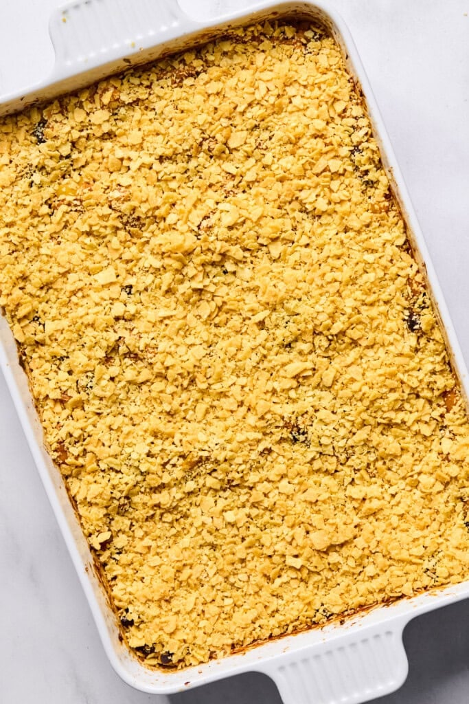 A white baking dish filled with a Vegan Mexican Casserole, topped with a layer of crushed yellow crackers or chips, sits on a marble surface.