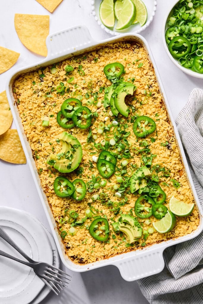 A Vegan Mexican Casserole topped with crushed chips, sliced avocados, jalapeños, green onions, and lime wedges sits on a white table with tortilla chips, lime slices, and green onions nearby.