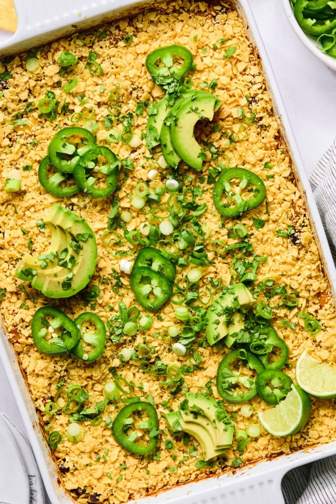 A Vegan Mexican Casserole baked to perfection, topped with crushed chips, sliced avocado, jalapeño, green onions, and lime wedges, displayed in a white dish with a spoon and a small bowl of greens nearby.