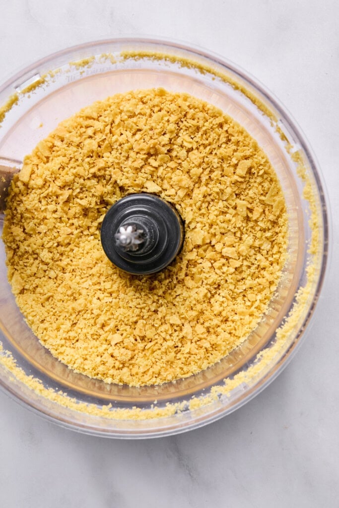 A food processor bowl filled with finely crushed yellow cornflakes, ideal for topping a Vegan Mexican Casserole, shown from above on a light surface.