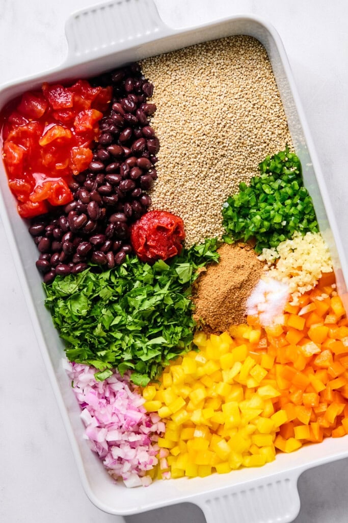 Rectangular dish with separate piles of diced tomatoes, black beans, quinoa, tomato paste, chopped cilantro, diced jalapeños, chili powder, salt, chopped red onion, orange and yellow bell peppers—perfect for assembling a flavorful Vegan Mexican Casserole.