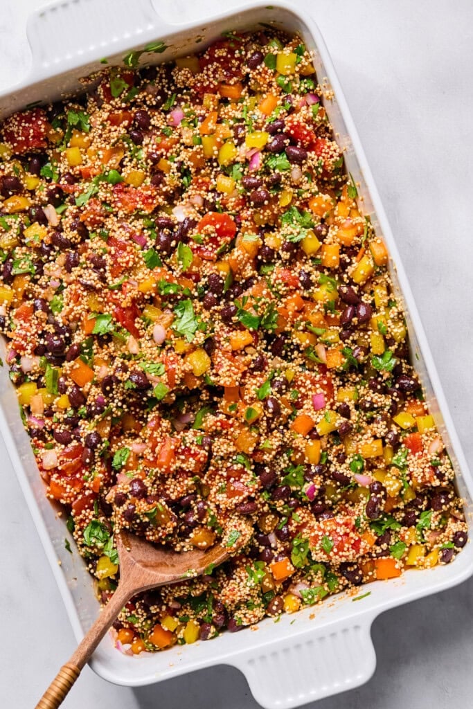 Colorful Vegan Mexican Casserole featuring quinoa, black beans, chopped red, yellow, and orange bell peppers, tomatoes, onions, and herbs in a white baking dish. A wooden spoon rests inside, ready for serving.