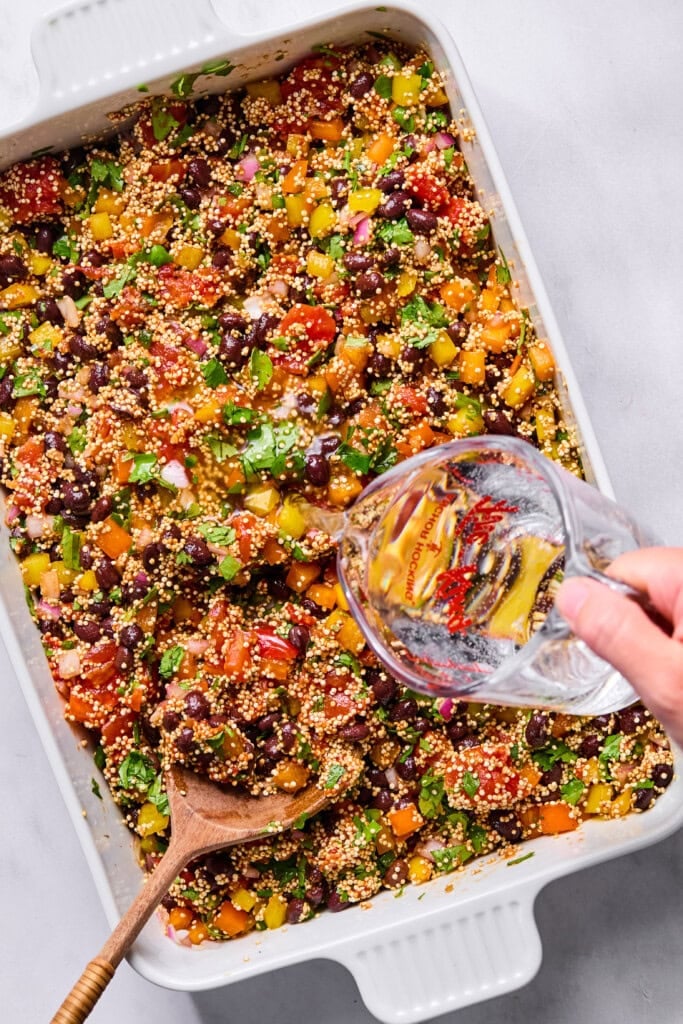 A hand pours water from a glass pitcher into a baking dish filled with a colorful mixture of quinoa, beans, chopped vegetables, and herbs, creating the base for a delicious Vegan Mexican Casserole. A wooden spoon is resting in the dish.