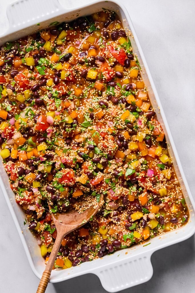 A white baking dish filled with a colorful mixture of chopped vegetables, black beans, quinoa, and herbs—this vibrant Vegan Mexican Casserole comes together beautifully, with a wooden spoon resting inside.