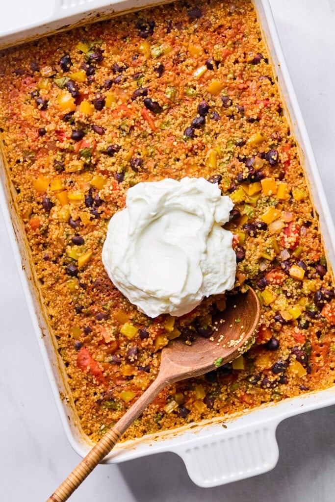 A Vegan Mexican Casserole featuring baked quinoa and vegetables sits in a white dish, topped with a dollop of sour cream. A wooden spoon rests inside, ready to serve.