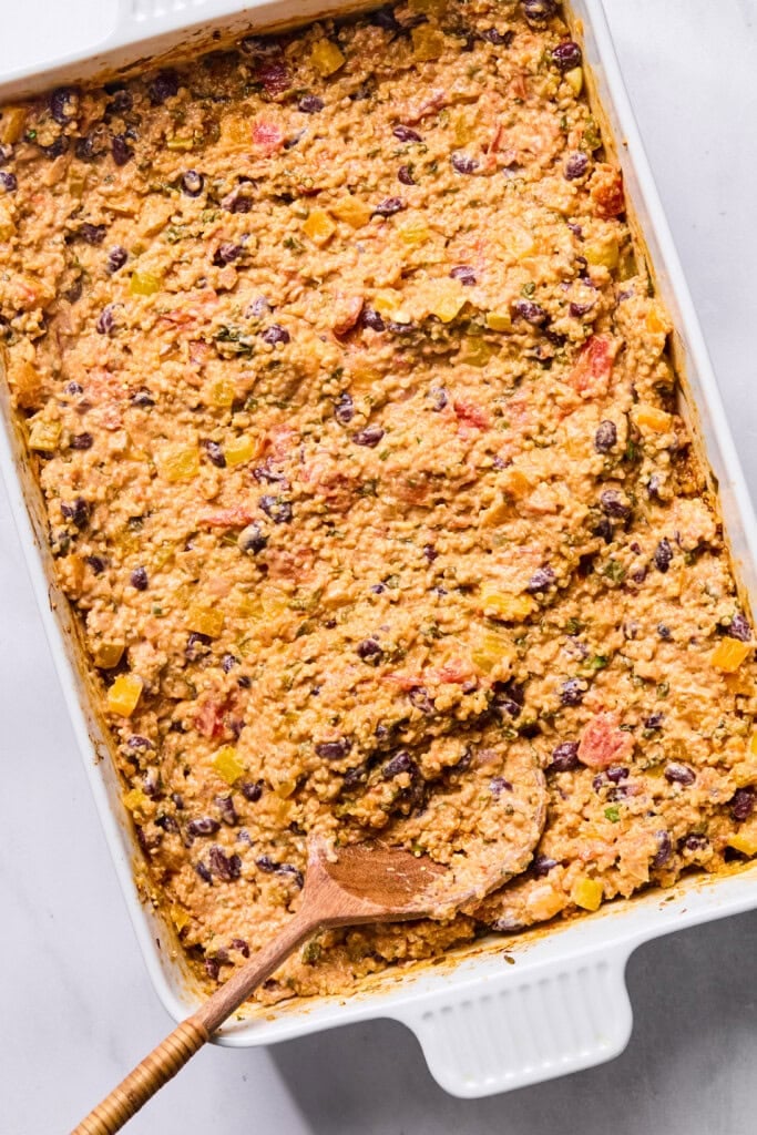 A baking dish filled with a textured, mixed Vegan Mexican Casserole of beans, grains, and vegetables, with a wooden spoon scooping some from the corner.