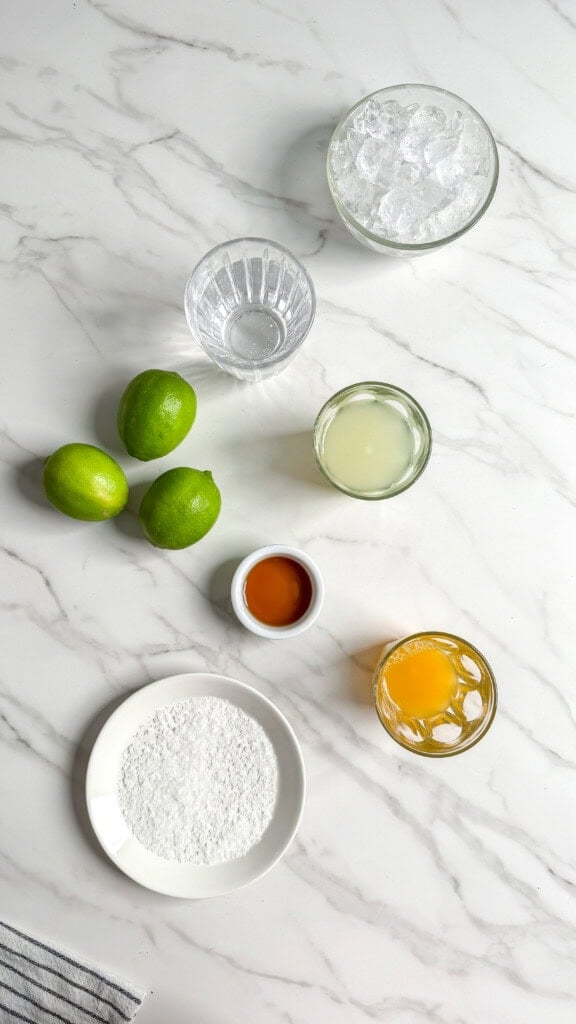 Overhead view of Fresh Classic Margaritas ingredients on a marble surface: a bowl of ice, three whole limes, a bowl of lime juice, a small bowl of brown liquid, powdered sugar, a glass of orange liquid, and an empty glass.