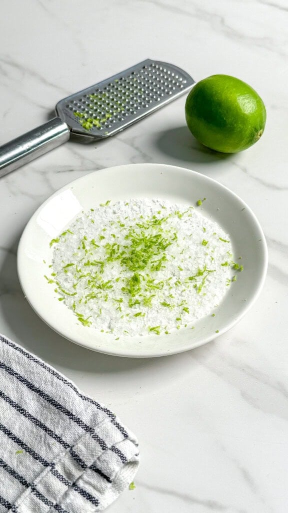 A white plate with coarse salt and lime zest—perfect for rimming Fresh Classic Margaritas—sits on a marble surface beside a whole lime, metal grater, and a striped dish towel peeking in the corner.