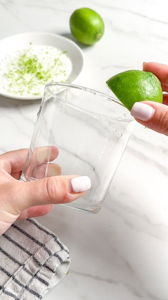 A person rubs a lime wedge on the rim of a clear glass, preparing for Fresh Classic Margaritas, with a plate of lime zest and a whole lime in the background on a white surface.